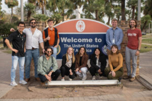 Group of 10 smiling law students, five crouched, five standing, wearing casual clothing pose in front of an orange and blue sign that reads "Welcome to The United States of America". This sign is located on Texas Southmost College campus, and faces the International Bridge with Mexico.