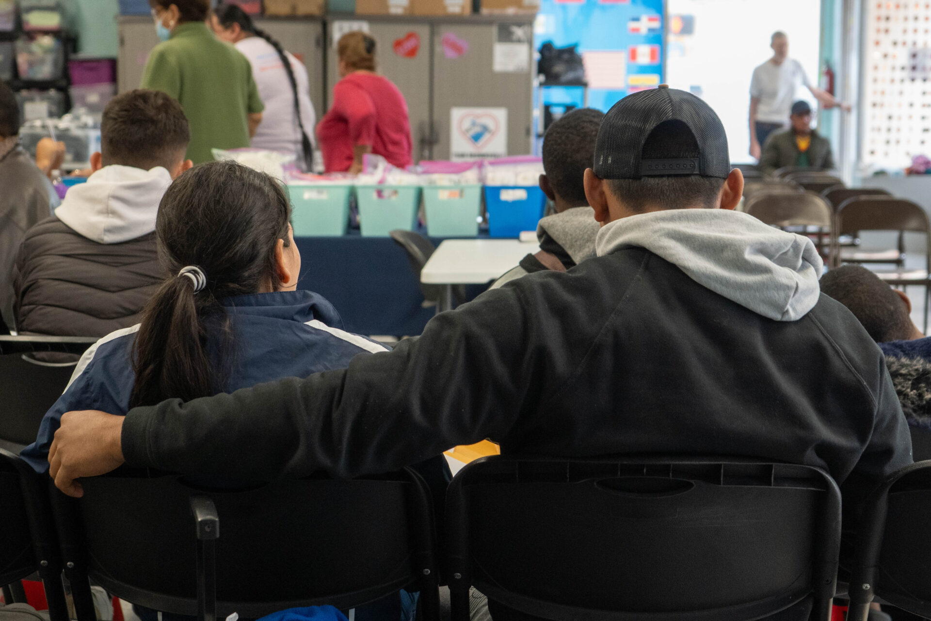 A young man and woman sit and wait patiently to fill out a Work Authorization application. They are wearing darkly colored jackets, the young man is wearing a baseball cap, the young woman has her hair in a ponytail, and the young man has his arm cradled along the back of the young woman's chair.