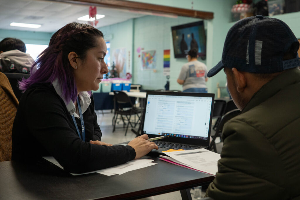 A ProBAR team member with a black sweater and long black hair styled behind her ears assists a man in a dark green jacket and a baseball cap with his Work Authorization application. The camera faces the computer between them (screen blurred), beyond them, the colorful, welcoming decor of the Good Neighbor Settlement House facility can be seen.