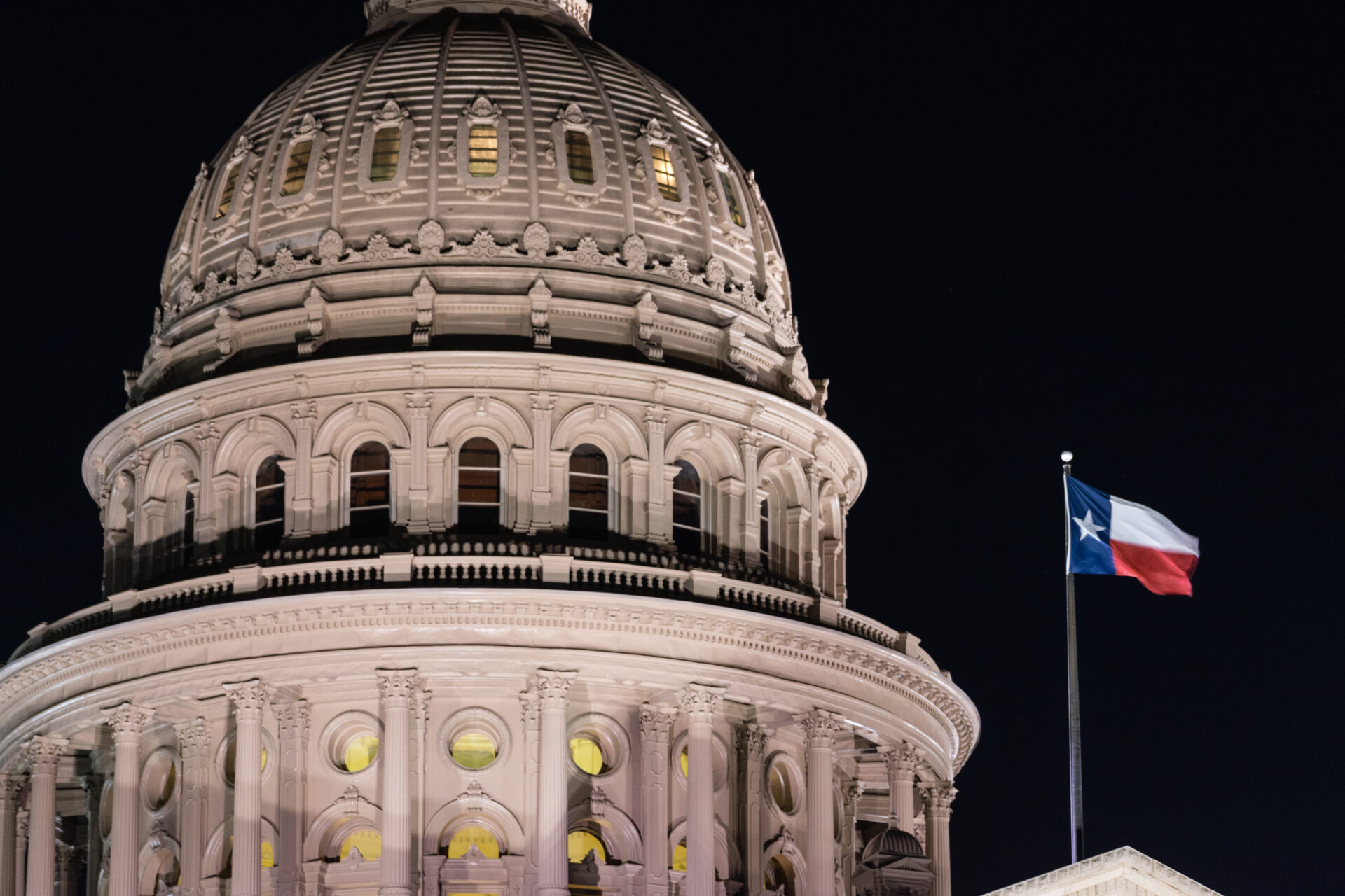A striking photo of the dome of the Texas State Capitol building at night, lit brightly from below. Standing independently of the dome is a flag pole with the Texas state flag at the top, also lit from below, creating a stark contrast to the darkness of the night sky.