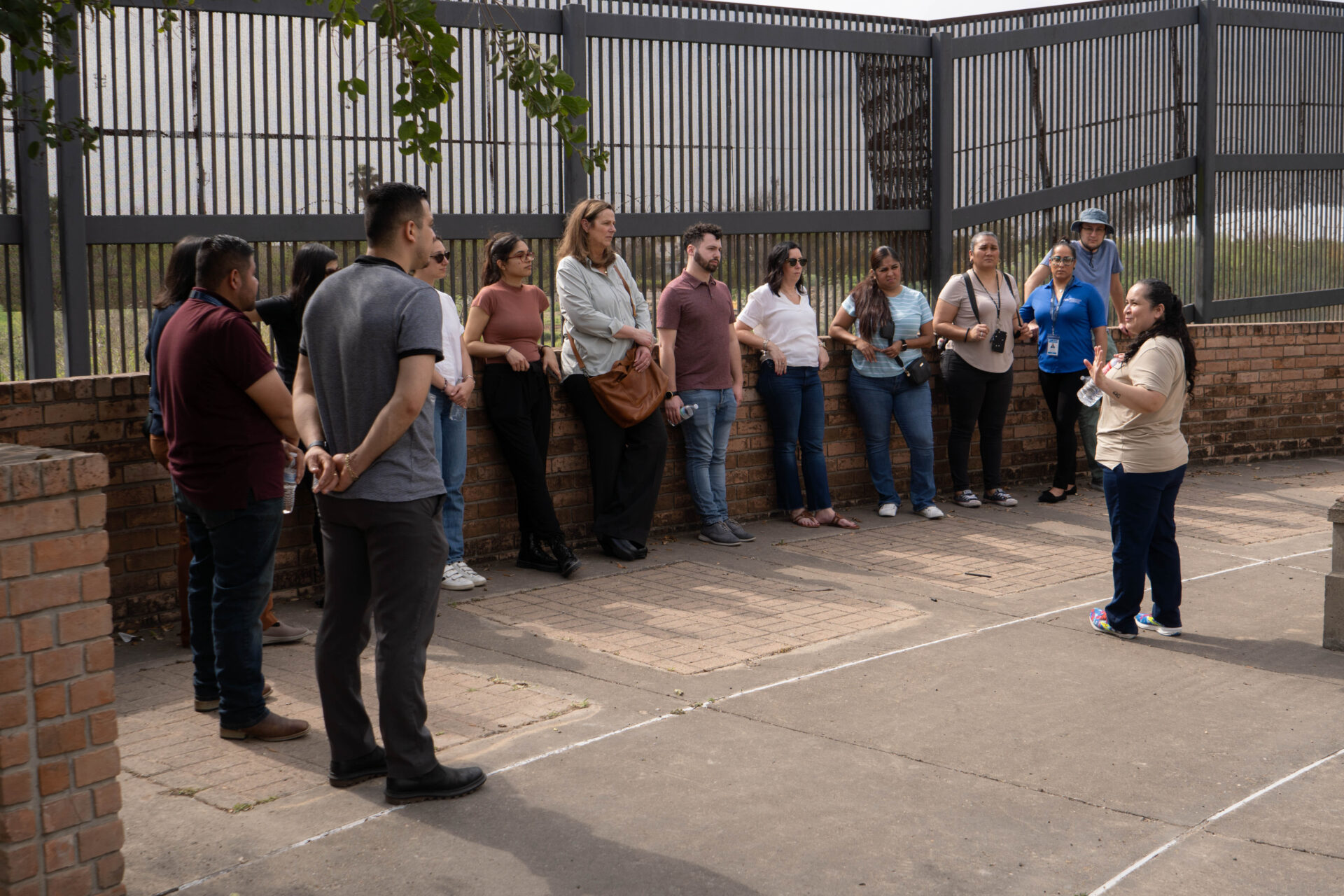 A group of law students listens intently to a presenter while on a tour of the border fence in Brownsville, Texas. The fence stands tall behind them.