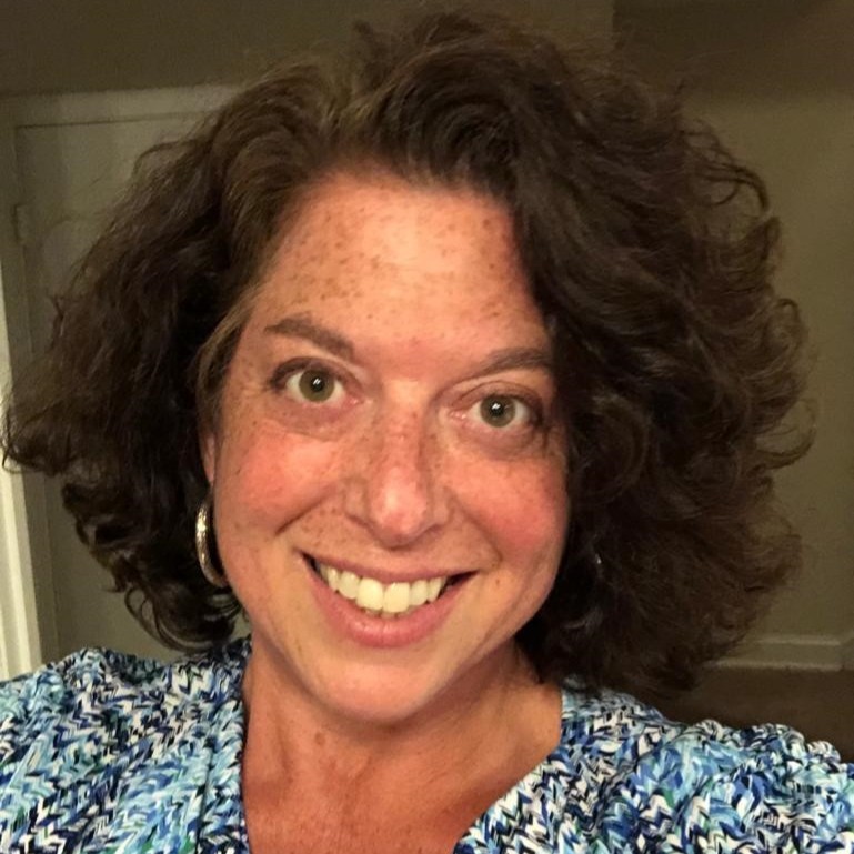 a tan woman with freckles and a big smile, medium length flowing dark brown hair, wearing gold hoop earrings and a floral blue blouse