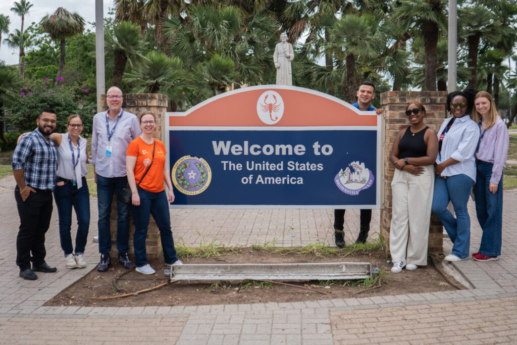 group of men and women of varying skin tones and heights standing and smiling in front of an orange and blue sign that reads 'Welcome to the United States of America'