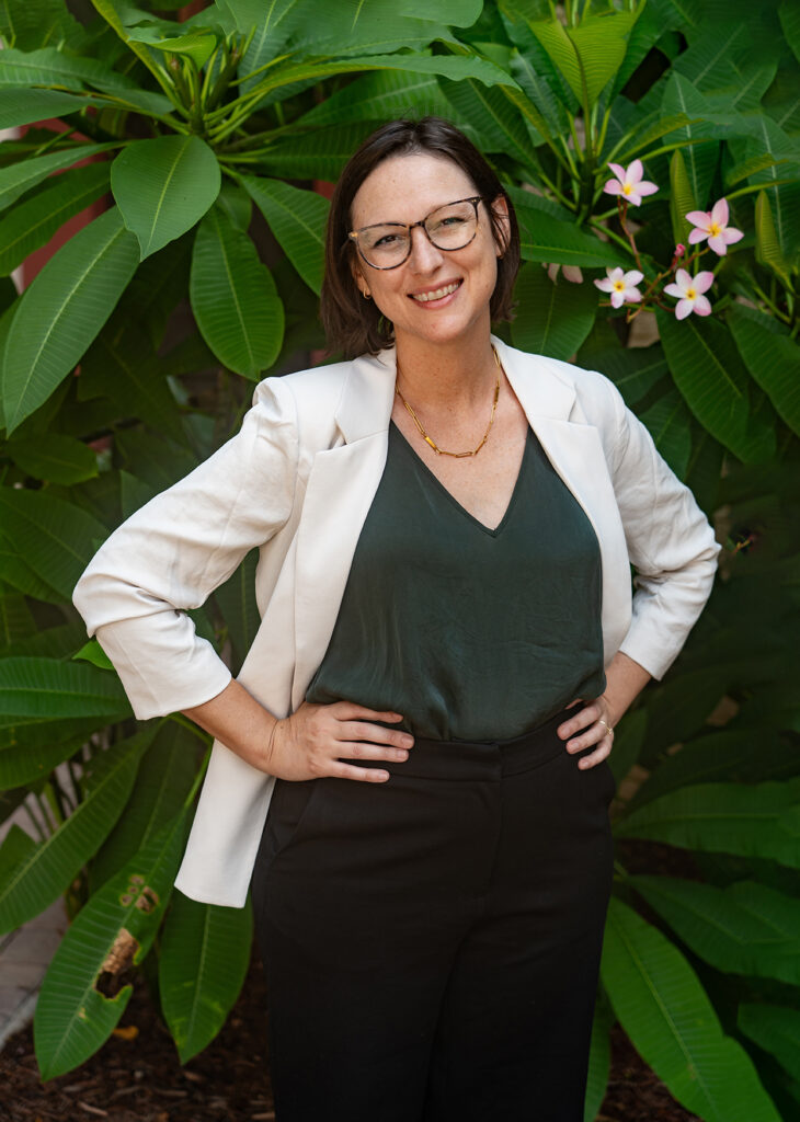 Photo of a smiling caucasian woman wearing glasses, shoulder length hair, and a white blazer with her hands on her hips. Standing in front of a bright green tropical plant.